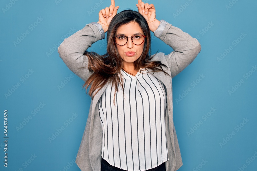 Young hispanic business woman wearing glasses standing over blue isolated background doing funny gesture with finger over head as bull horns Young hispanic business woman wearing glasses standing over blue isolated background doing funny gesture with finger over head as bull horns