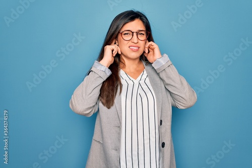Young hispanic business woman wearing glasses standing over blue isolated background covering ears with fingers with annoyed expression for the noise of loud music. Deaf concept.