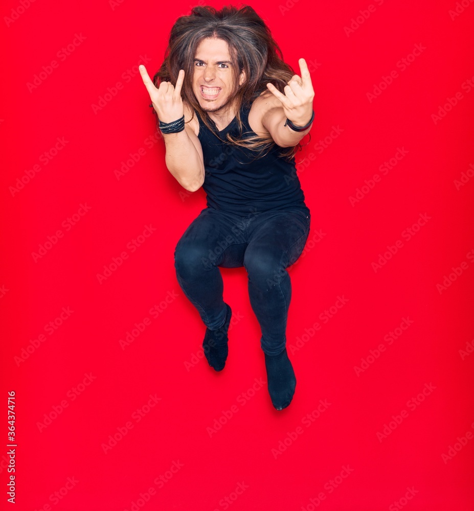 Young handsome man with long hair with aggressive expression wearing casual clothes . Jumping doing heavy metal sign over isolated red background.