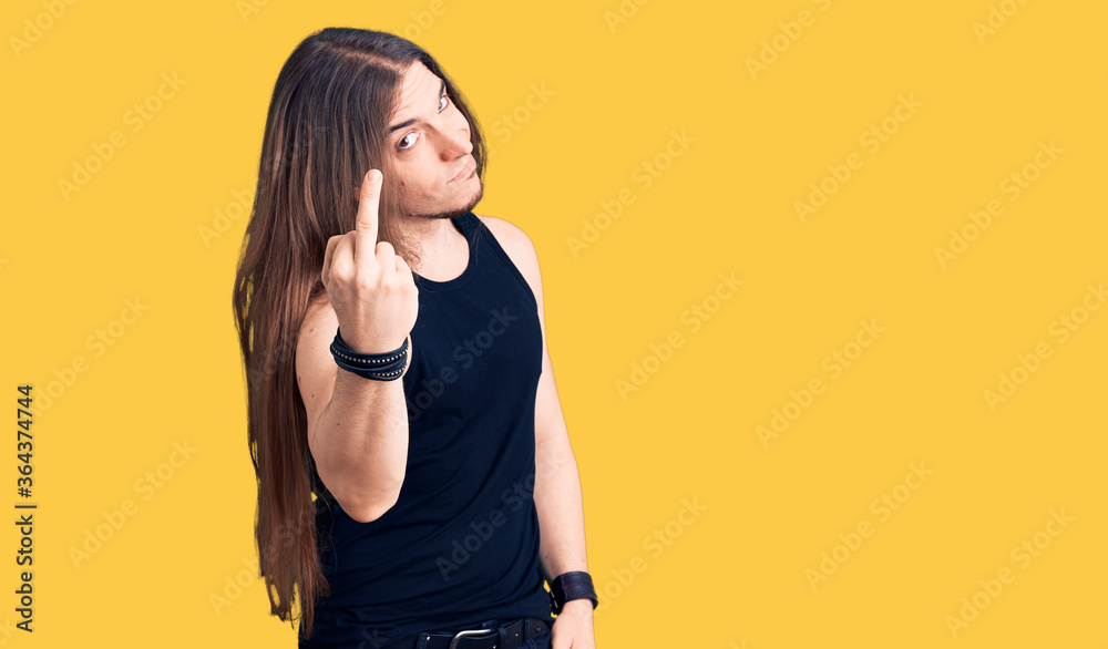 Young adult man with long hair wearing goth style with black clothes showing middle finger, impolite and rude fuck off expression Young adult man with long hair wearing goth style with black clothes showing middle finger, impolite and rude fuck off expression