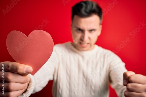 Young handsome caucasian man holding paper shape heart over red background annoyed and frustrated shouting with anger, crazy and yelling with raised hand, anger concept © Krakenimages.com