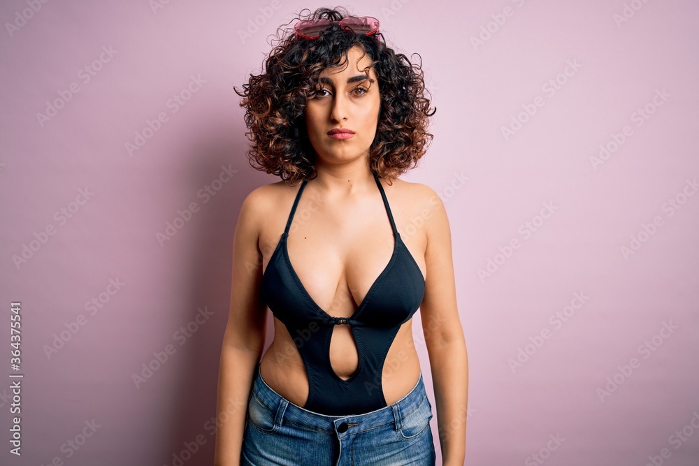 Young beautiful arab woman on vacation wearing swimsuit and sunglasses over pink background with serious expression on face. Simple and natural looking at the camera. Young beautiful arab woman on vacation wearing swimsuit and sunglasses over pink background with serious expression on face. Simple and natural looking at the camera.