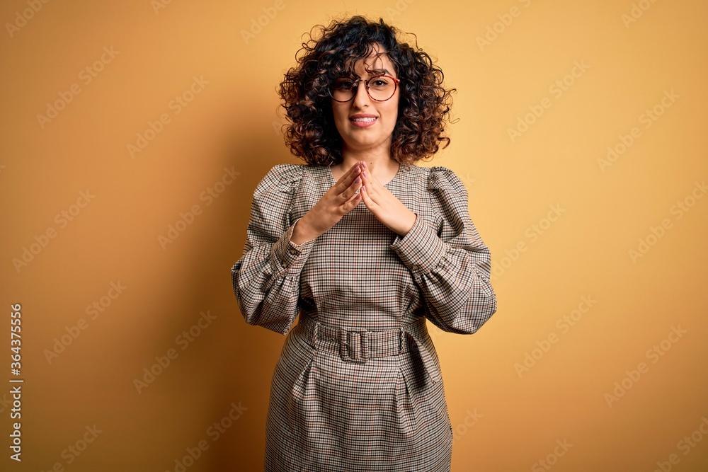 Beautiful arab business woman wearing dress and glasses standing over yellow background Hands together and fingers crossed smiling relaxed and cheerful. Success and optimistic Beautiful arab business woman wearing dress and glasses standing over yellow background Hands together and fingers crossed smiling relaxed and cheerful. Success and optimistic