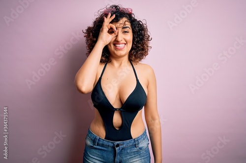 Young beautiful arab woman on vacation wearing swimsuit and sunglasses over pink background doing ok gesture with hand smiling, eye looking through fingers with happy face.