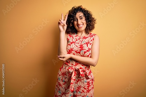 Young beautiful curly arab woman on vacation wearing summer floral dress and sunglasses smiling with happy face winking at the camera doing victory sign. Number two.