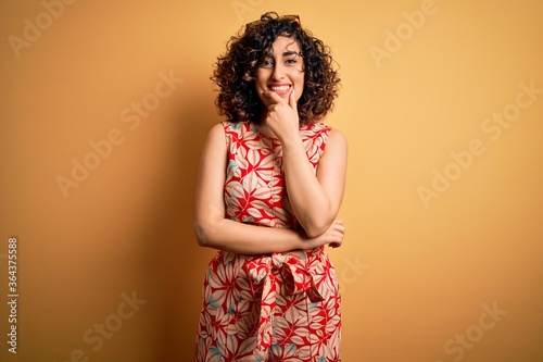 Young beautiful curly arab woman on vacation wearing summer floral dress and sunglasses looking confident at the camera smiling with crossed arms and hand raised on chin. Thinking positive.