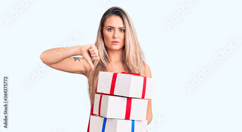 Young beautiful blonde woman holding birthday gifts with angry face  negative sign showing dislike with thumbs down  rejection concept