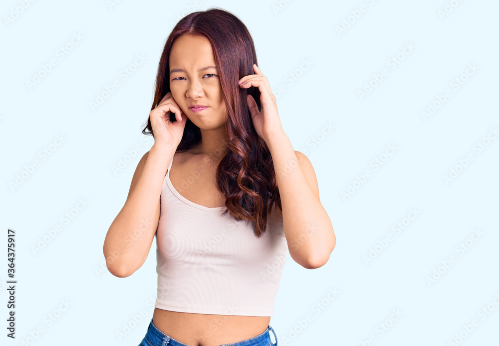 Young beautiful chinese girl wearing casual clothes covering ears with fingers with annoyed expression for the noise of loud music. deaf concept.