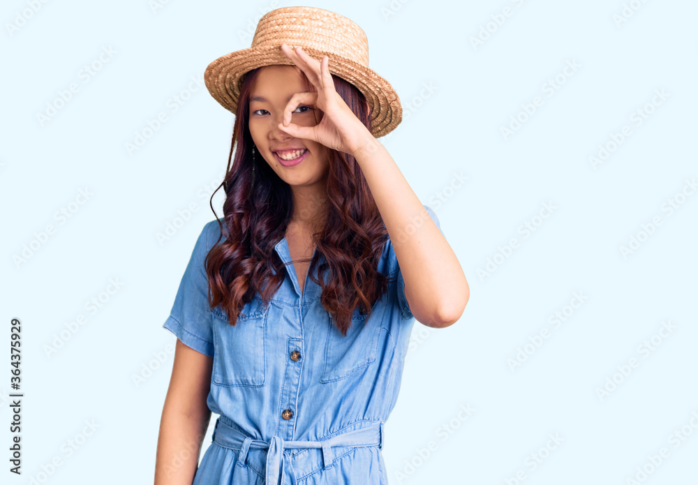 Young beautiful chinese girl wearing summer hat doing ok gesture with hand smiling, eye looking through fingers with happy face. Young beautiful chinese girl wearing summer hat doing ok gesture with hand smiling, eye looking through fingers with happy face.