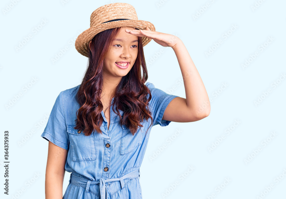 Young beautiful chinese girl wearing summer hat very happy and smiling looking far away with hand over head. searching concept. Young beautiful chinese girl wearing summer hat very happy and smiling looking far away with hand over head. searching concept.