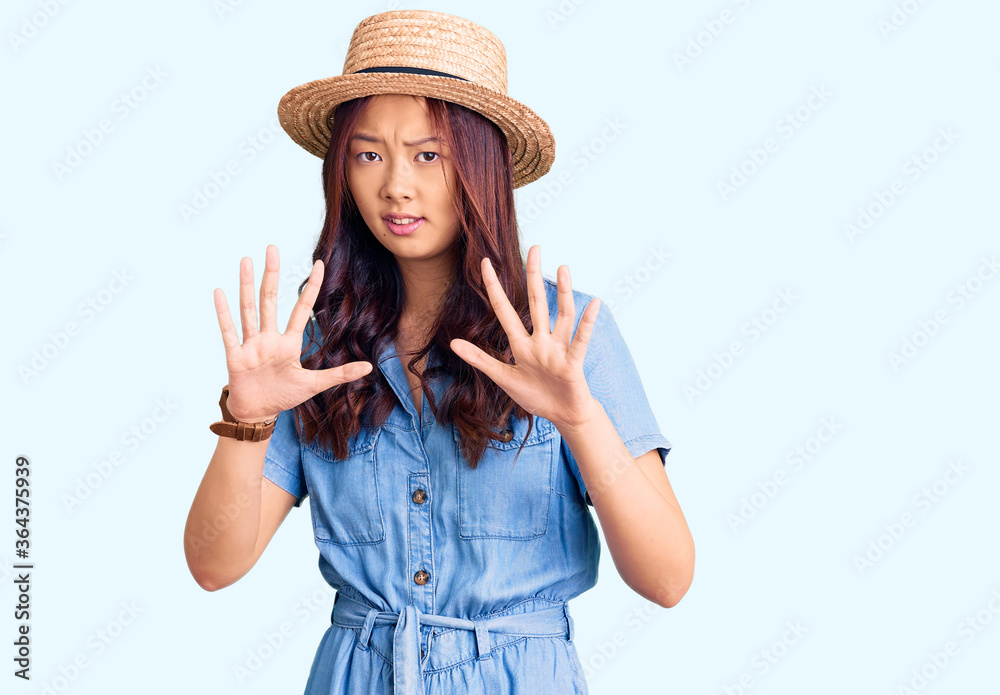 Young beautiful chinese girl wearing summer hat afraid and terrified with fear expression stop gesture with hands, shouting in shock. panic concept. Young beautiful chinese girl wearing summer hat afraid and terrified with fear expression stop gesture with hands, shouting in shock. panic concept.