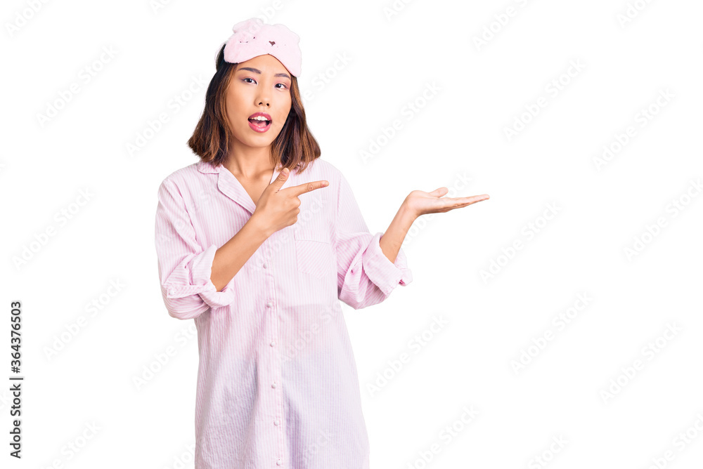 Young beautiful chinese girl wearing sleep mask and pajama amazed and smiling to the camera while presenting with hand and pointing with finger. Young beautiful chinese girl wearing sleep mask and pajama amazed and smiling to the camera while presenting with hand and pointing with finger.