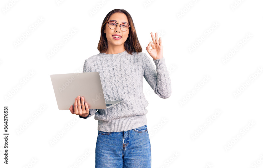 Young beautiful chinese girl wearing glasses holding laptop doing ok sign with fingers, smiling friendly gesturing excellent symbol Young beautiful chinese girl wearing glasses holding laptop doing ok sign with fingers, smiling friendly gesturing excellent symbol