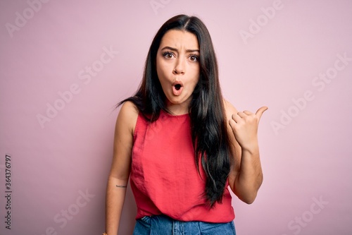 Young brunette woman wearing casual summer shirt over pink isolated background Surprised pointing with hand finger to the side, open mouth amazed expression.