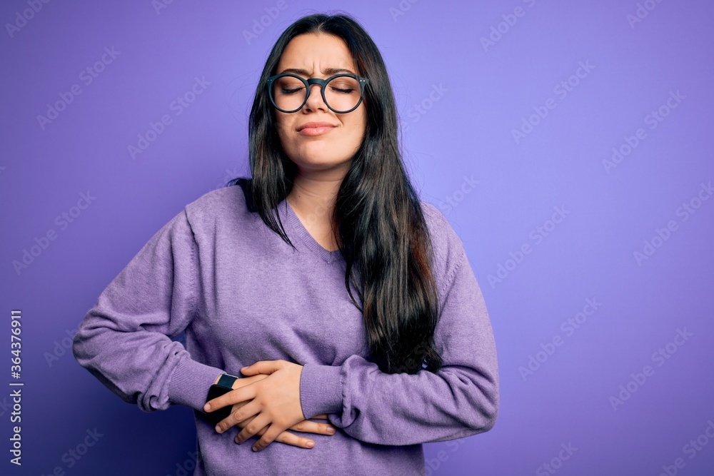 Young brunette woman wearing glasses over purple isolated background with hand on stomach because indigestion, painful illness feeling unwell. Ache concept.