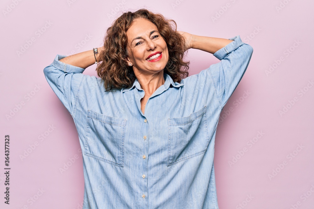 Middle age beautiful woman wearing casual denim shirt standing over pink background relaxing and stretching, arms and hands behind head and neck smiling happy Middle age beautiful woman wearing casual denim shirt standing over pink background relaxing and stretching, arms and hands behind head and neck smiling happy