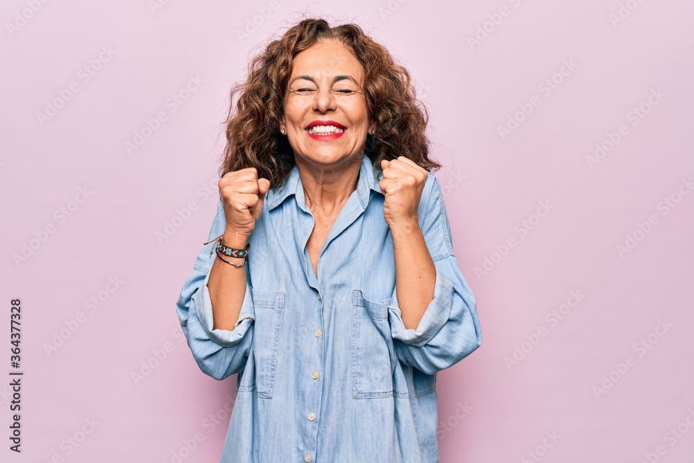 Middle age beautiful woman wearing casual denim shirt standing over pink background excited for success with arms raised and eyes closed celebrating victory smiling. Winner concept. Middle age beautiful woman wearing casual denim shirt standing over pink background excited for success with arms raised and eyes closed celebrating victory smiling. Winner concept.