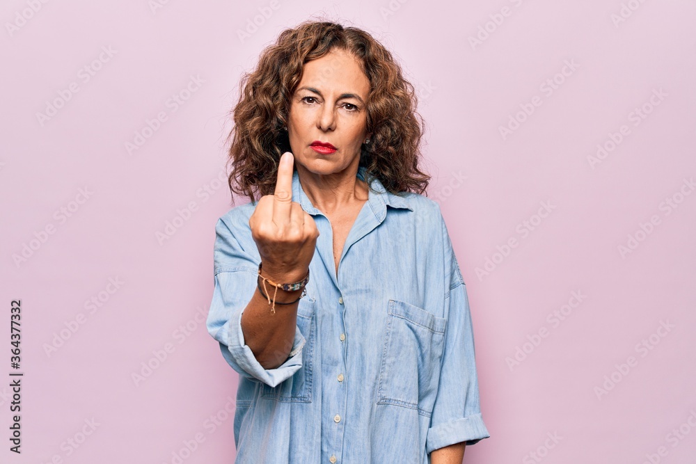 Middle age beautiful woman wearing casual denim shirt standing over pink background Showing middle finger, impolite and rude fuck off expression Middle age beautiful woman wearing casual denim shirt standing over pink background Showing middle finger, impolite and rude fuck off expression