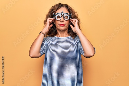Middle age beautiful woman controlling vision using optometry glasses over yellow background with hand on head  headache because stress. Suffering migraine.