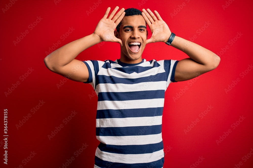 Handsome african american man wearing casual striped t-shirt standing over red background Smiling cheerful playing peek a boo with hands showing face. Surprised and exited Handsome african american man wearing casual striped t-shirt standing over red background Smiling cheerful playing peek a boo with hands showing face. Surprised and exited