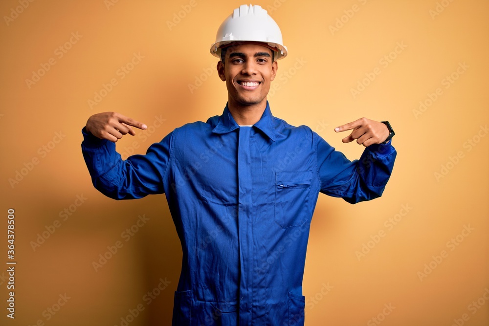 Young handsome african american worker man wearing blue uniform and security helmet looking confident with smile on face, pointing oneself with fingers proud and happy.