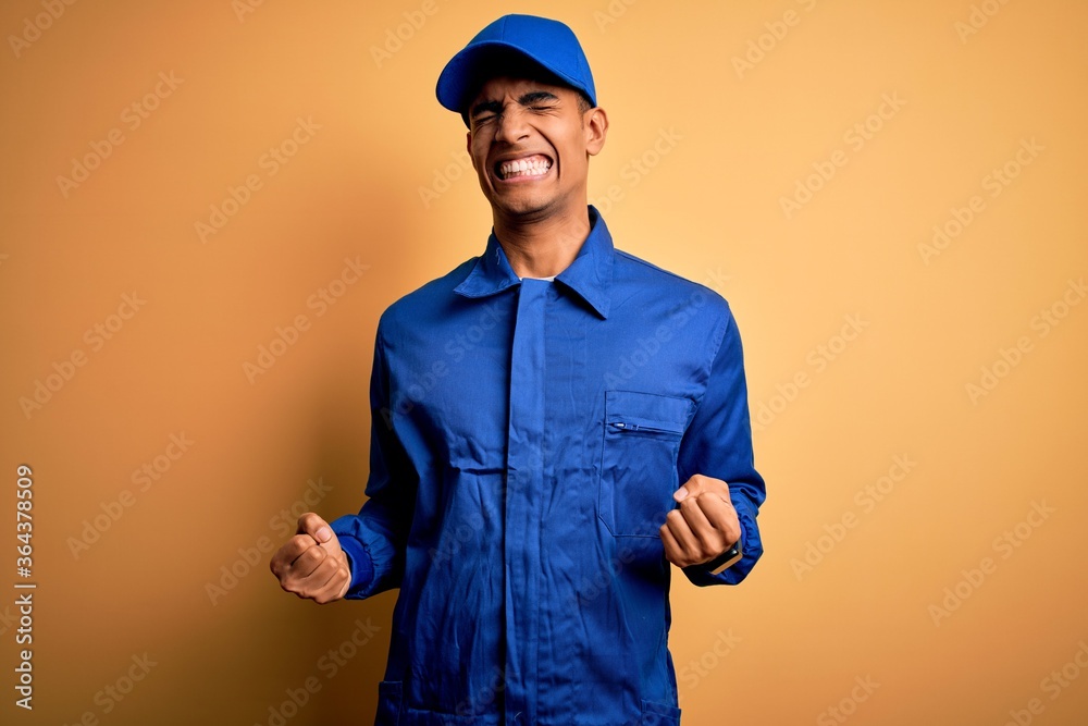 Young african american mechanic man wearing blue uniform and cap over yellow background very happy and excited doing winner gesture with arms raised, smiling and screaming for success. Celebration. Young african american mechanic man wearing blue uniform and cap over yellow background very happy and excited doing winner gesture with arms raised, smiling and screaming for success. Celebration.