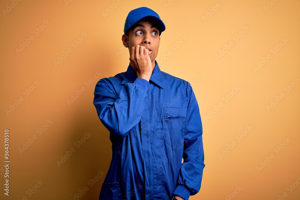 Young african american mechanic man wearing blue uniform and cap over yellow background looking stressed and nervous with hands on mouth biting nails. Anxiety problem.