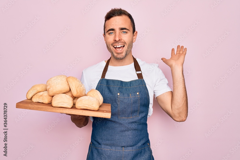 Young handsome baker man with blue eyes wearing apron holding tray with homemade bread very happy and excited, winner expression celebrating victory screaming with big smile and raised hands Young handsome baker man with blue eyes wearing apron holding tray with homemade bread very happy and excited, winner expression celebrating victory screaming with big smile and raised hands