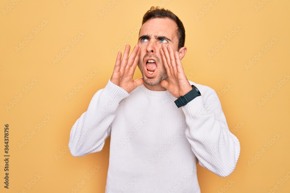 Young handsome man with blue eyes wearing casual sweater standing over yellow background Shouting angry out loud with hands over mouth Young handsome man with blue eyes wearing casual sweater standing over yellow background Shouting angry out loud with hands over mouth