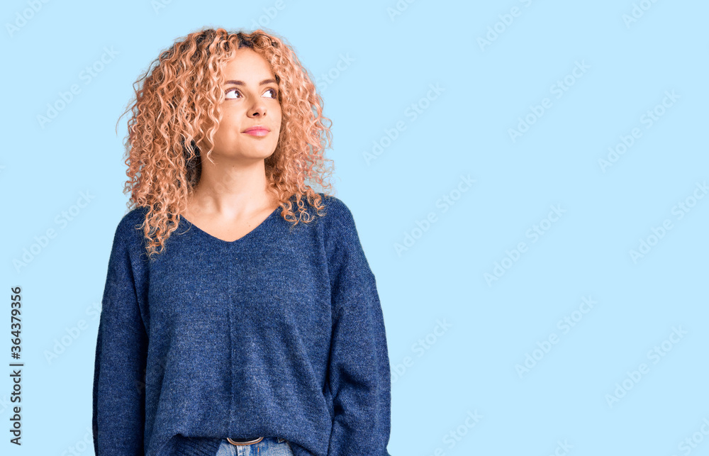 Young blonde woman with curly hair wearing casual winter sweater smiling looking to the side and staring away thinking. Young blonde woman with curly hair wearing casual winter sweater smiling looking to the side and staring away thinking.