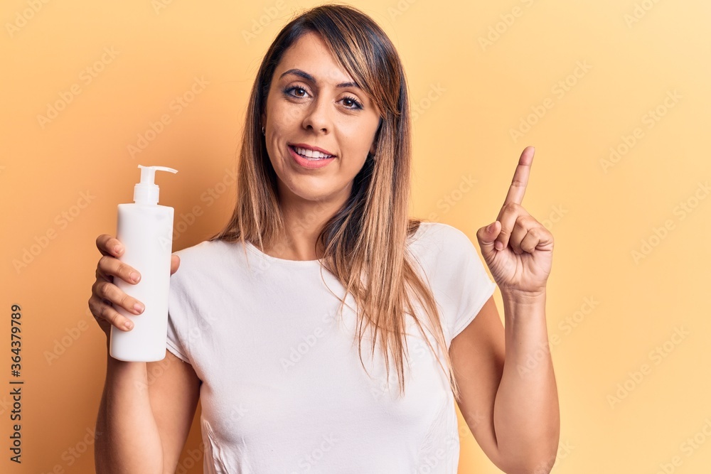 Young beautiful woman holding bottle of cream lotion smiling happy pointing with hand and finger to the side Young beautiful woman holding bottle of cream lotion smiling happy pointing with hand and finger to the side