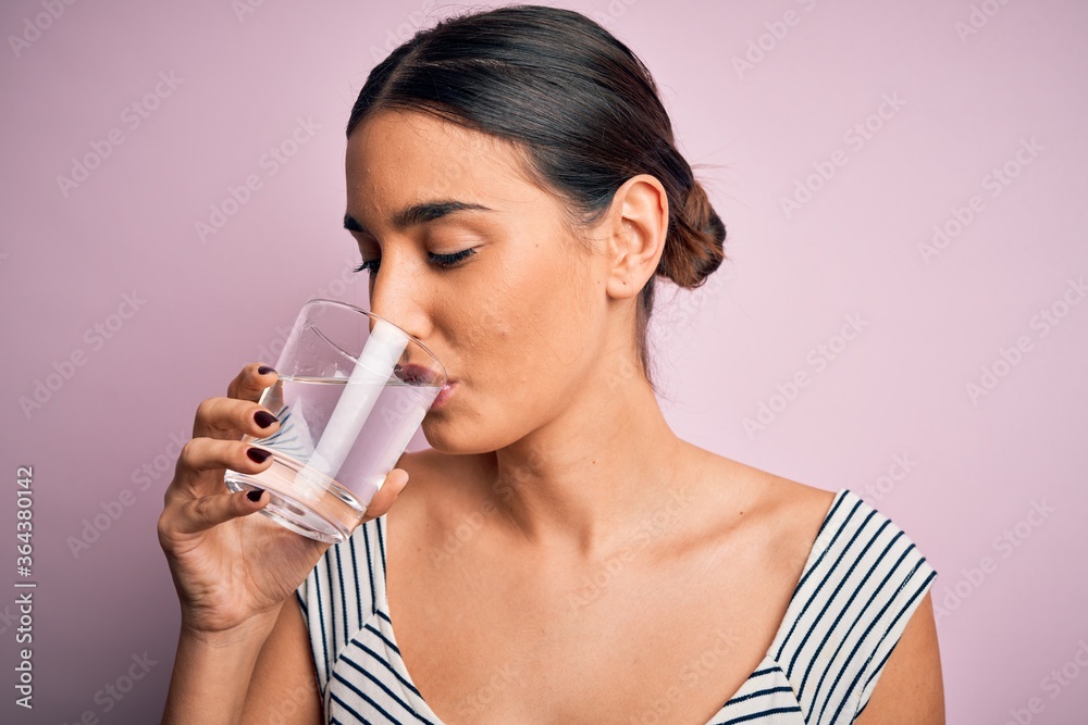 Young beautiful brunette woman drinking glass of healthy water to refreshment standing over isolated pink background Young beautiful brunette woman drinking glass of healthy water to refreshment standing over isolated pink background