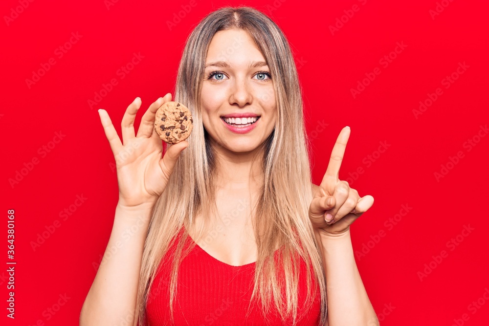 Young beautiful blonde woman holding cookie smiling with an idea or question pointing finger with happy face, number one Young beautiful blonde woman holding cookie smiling with an idea or question pointing finger with happy face, number one