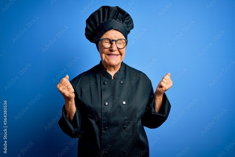 Senior beautiful grey-haired chef woman wearing cooker uniform and hat over blue background very happy and excited doing winner gesture with arms raised, smiling and screaming for success. Celebration Senior beautiful grey-haired chef woman wearing cooker uniform and hat over blue background very happy and excited doing winner gesture with arms raised, smiling and screaming for success. Celebration