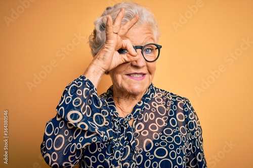 Senior beautiful grey-haired woman wearing casual shirt and glasses over yellow background doing ok gesture with hand smiling, eye looking through fingers with happy face.