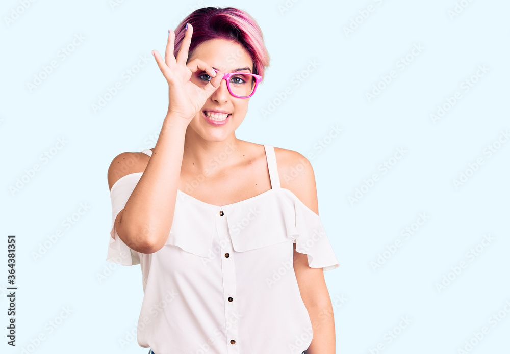 Young beautiful woman with pink hair wearing casual clothes and glasses doing ok gesture with hand smiling, eye looking through fingers with happy face.
