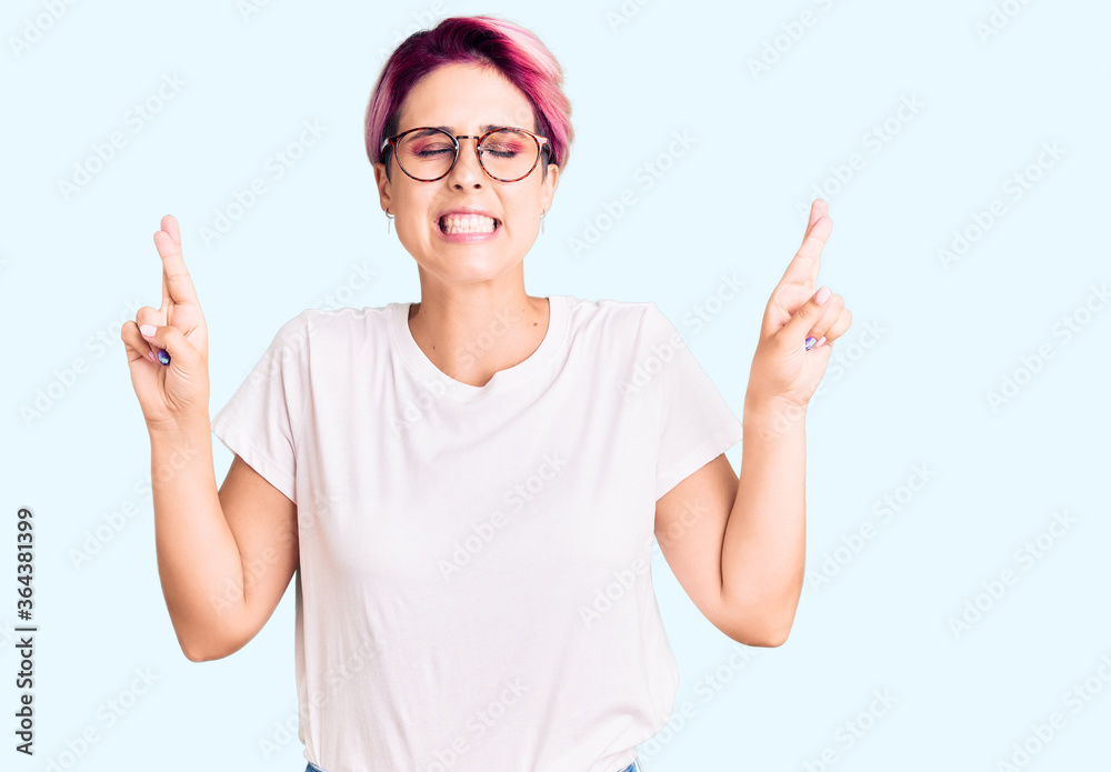 Young beautiful woman with pink hair wearing casual clothes and glasses gesturing finger crossed smiling with hope and eyes closed. luck and superstitious concept. Young beautiful woman with pink hair wearing casual clothes and glasses gesturing finger crossed smiling with hope and eyes closed. luck and superstitious concept.