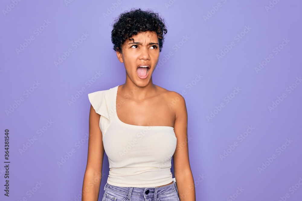 Young beautiful african american afro woman wearing casual t-shirt over purple background angry and mad screaming frustrated and furious, shouting with anger. Rage and aggressive concept.
