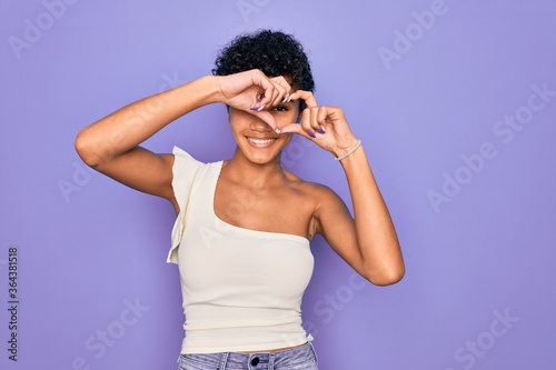 Young beautiful african american afro woman wearing casual t-shirt over purple background Doing heart shape with hand and fingers smiling looking through sign