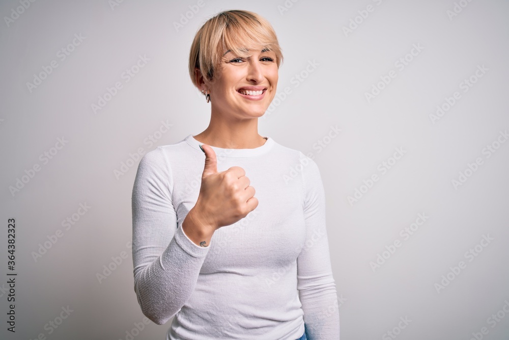 Young beautiful blonde woman with modern short hair hairstyle standing over isolated background doing happy thumbs up gesture with hand. Approving expression looking at the camera showing success. Young beautiful blonde woman with modern short hair hairstyle standing over isolated background doing happy thumbs up gesture with hand. Approving expression looking at the camera showing success.