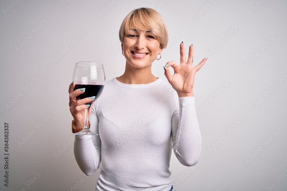 Young blonde woman with short hair drinking a glass of red wine over isolated background doing ok sign with fingers, excellent symbol Young blonde woman with short hair drinking a glass of red wine over isolated background doing ok sign with fingers, excellent symbol