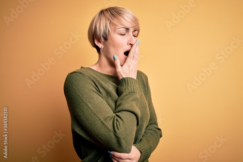 Young blonde woman with modern short hair wearing casual sweater over yellow background bored yawning tired covering mouth with hand. Restless and sleepiness.
