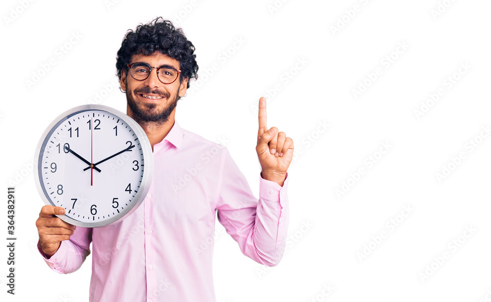 Handsome young man with curly hair and bear holding big clock surprised with an idea or question pointing finger with happy face, number one Handsome young man with curly hair and bear holding big clock surprised with an idea or question pointing finger with happy face, number one