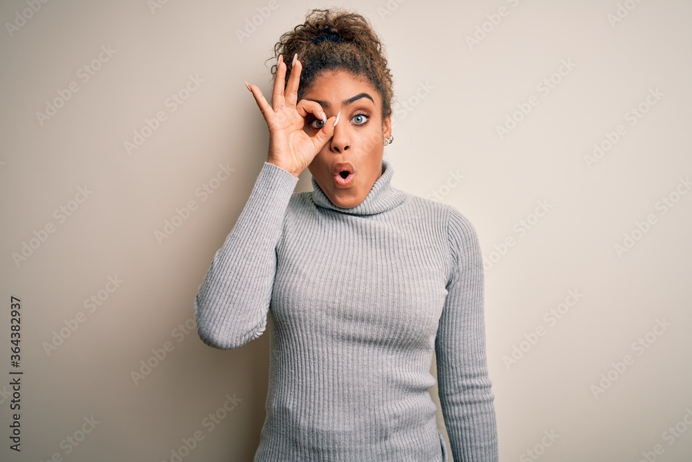 Beautiful african american girl wearing turtleneck sweater standing over white background doing ok gesture shocked with surprised face, eye looking through fingers. Unbelieving expression. Beautiful african american girl wearing turtleneck sweater standing over white background doing ok gesture shocked with surprised face, eye looking through fingers. Unbelieving expression.