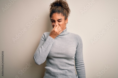Beautiful african american girl wearing turtleneck sweater standing over white background smelling something stinky and disgusting  intolerable smell  holding breath with fingers on nose. Bad smell