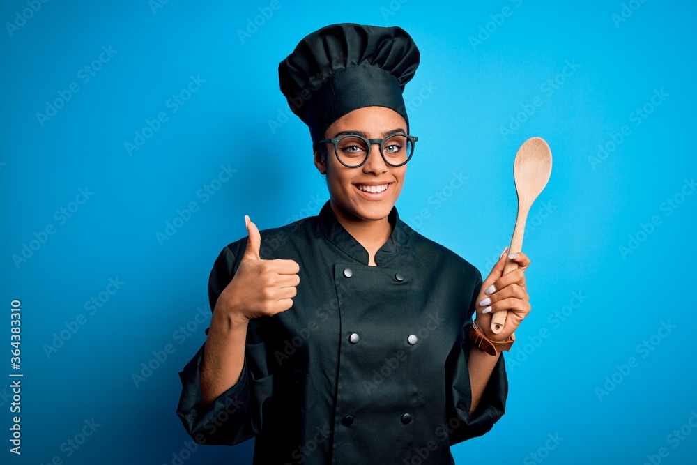 Fototapeta premium Young african american chef girl wearing cooker uniform and hat holding wooden spoon tool happy with big smile doing ok sign, thumb up with fingers, excellent sign