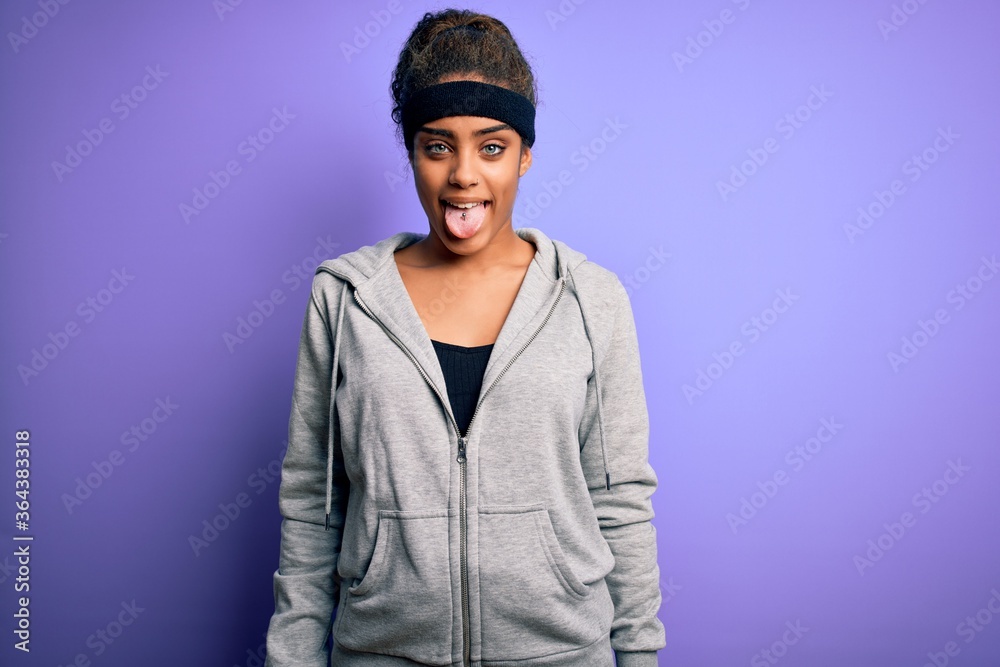Young african american sportswoman doing sport wearing sportswear over purple background sticking tongue out happy with funny expression. Emotion concept.