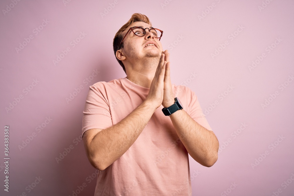Young handsome redhead man wearing casual t-shirt standing over isolated pink background begging and praying with hands together with hope expression on face very emotional and worried. Begging.