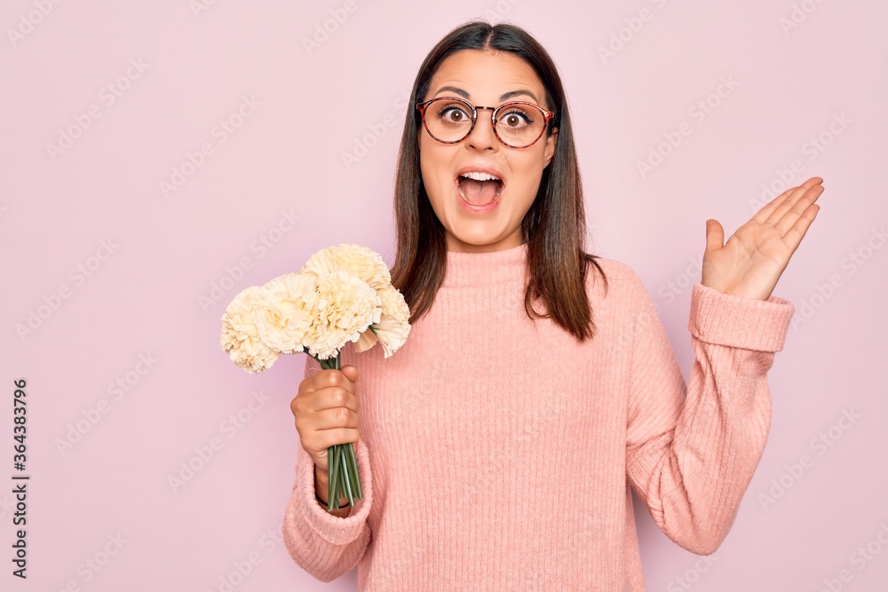 Beautiful brunette woman wearing glasses holding bouquet of flowers over pink background celebrating victory with happy smile and winner expression with raised hands Beautiful brunette woman wearing glasses holding bouquet of flowers over pink background celebrating victory with happy smile and winner expression with raised hands