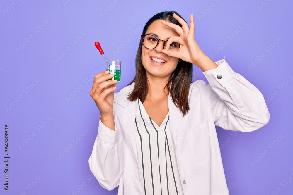 Young beautiful brunette scientist woman wearing coat and glasses holding test tube smiling happy doing ok sign with hand on eye looking through fingers Young beautiful brunette scientist woman wearing coat and glasses holding test tube smiling happy doing ok sign with hand on eye looking through fingers